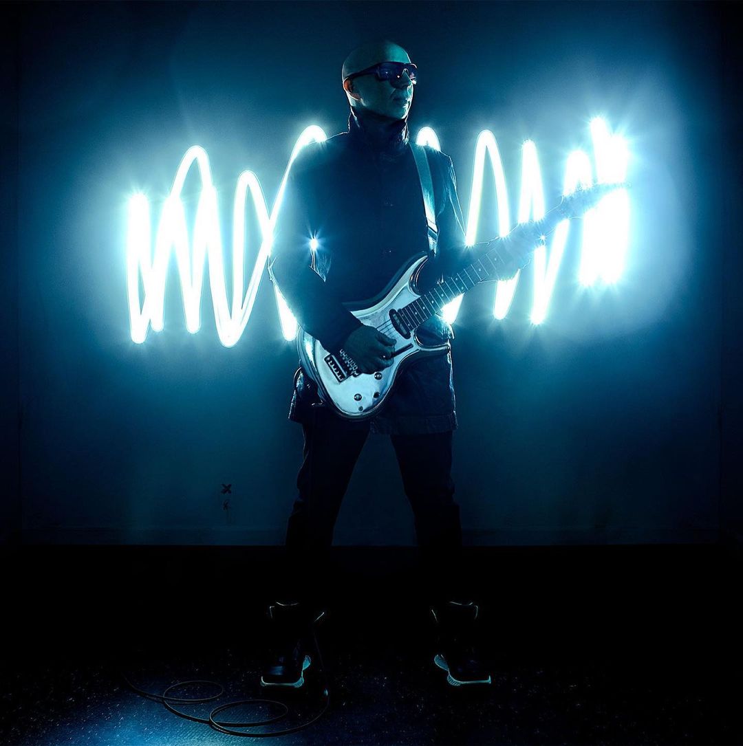 Man in black outfit playing white electric guitar on stage with neon squiggle lights behind him, bathed in blue light.