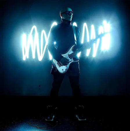 Man in black outfit playing white electric guitar on stage with neon squiggle lights behind him, bathed in blue light.