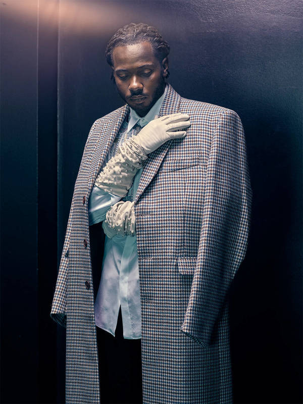 Portrait of a black male model in a patterned overcoat, blue dress shirt, and lace gloves posing in a dark indoor studio setting.