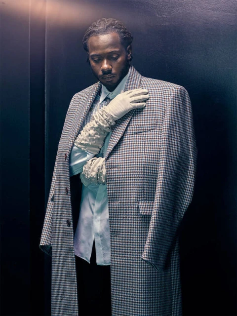 Portrait of a black male model in a patterned overcoat, blue dress shirt, and lace gloves posing in a dark indoor studio setting.