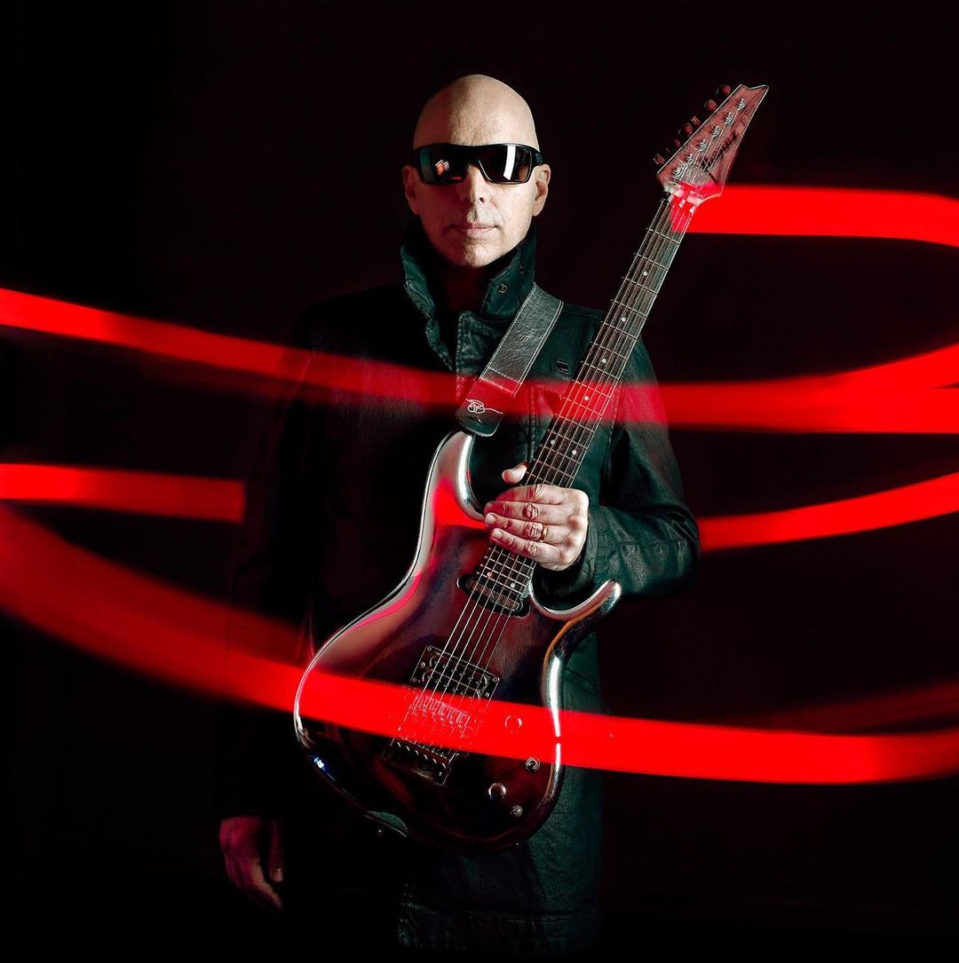 Bald man in sunglasses holding an electric guitar against a dark background with red light streaks, posing for a music-themed shot.