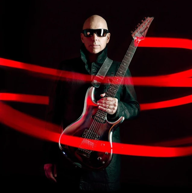 Bald man in sunglasses holding an electric guitar against a dark background with red light streaks, posing for a music-themed shot.