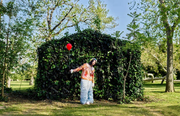 A woman in casual clothing with a red balloon stands in a grassy field next to a large green hedge with trees and blue sky overhead.
