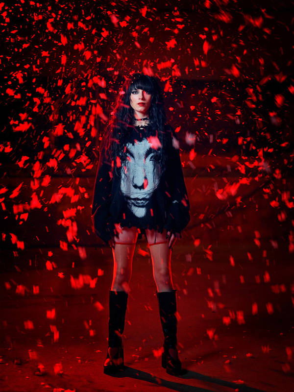 A woman with dark hair is illuminated by red lighting while wearing a black sweater with a face on it and black boots.