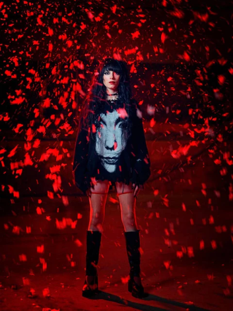 A woman with dark hair is illuminated by red lighting while wearing a black sweater with a face on it and black boots.