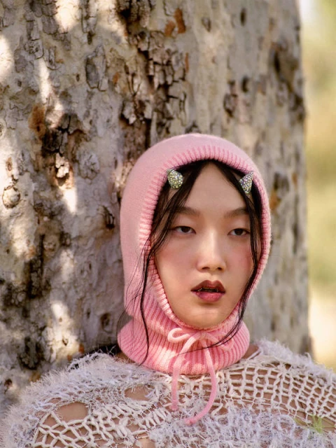 Asian female with a pink knit hood, face-framing strands, and jeweled hair clips, posing in front of a tree with dappled sunlight.