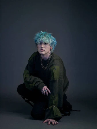 Portrait of a man with bright blue hair wearing an olive green sweater and layered chains against a grey studio background