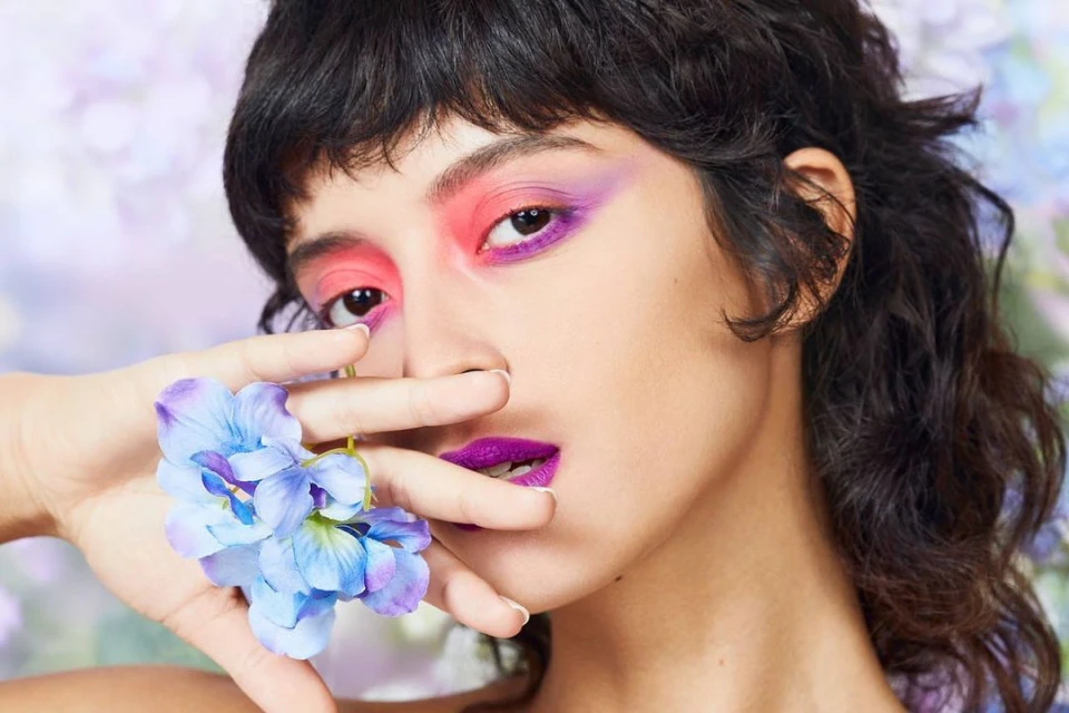 Close-up of a woman with vibrant pink and purple eye makeup, purple lipstick, and blue flowers near her face against a floral background.