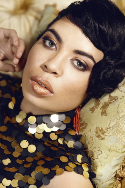 Close-up portrait of a woman with dark wavy hair and golden makeup, lying on a decorative pillow, wearing a sequined top.