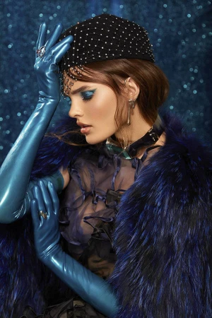 Model in blue latex gloves and feathered blue coat wearing a dark embellished cap against a sparkling blue background