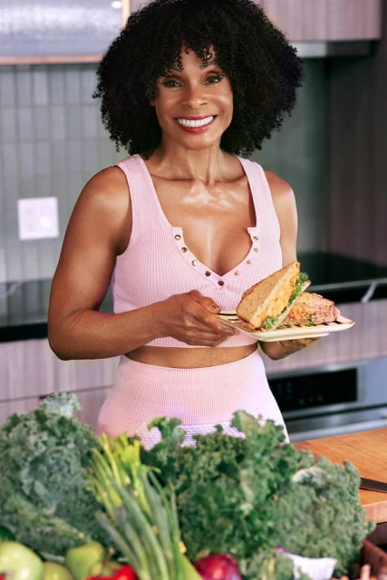 Adult female smiles while holding a sandwich in front of a kitchen counter filled with kale and other fruits and vegetables.