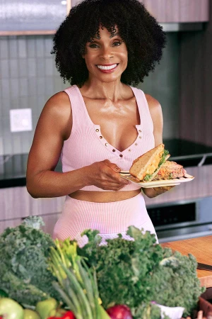 Adult female smiles while holding a sandwich in front of a kitchen counter filled with kale and other fruits and vegetables.