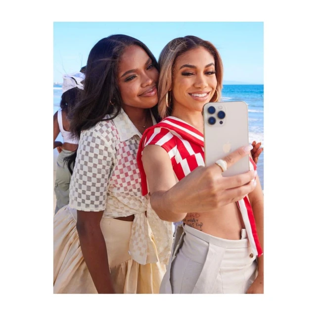 Two women pose for a beach selfie on a sunny day. One woman holds the phone, smiling, as her friend stands close beside her.