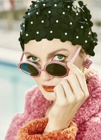 Woman wearing pink sunglasses, a textured pink coat, and a black floral swim cap with pearl centers, photographed by Nathalie Gordon.