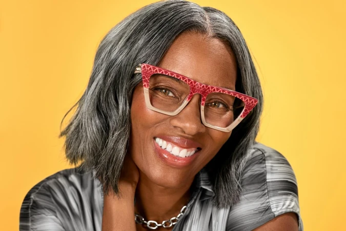 Portrait of an older black woman with grey hair wearing patterned red glasses and a grey top against a yellow background.