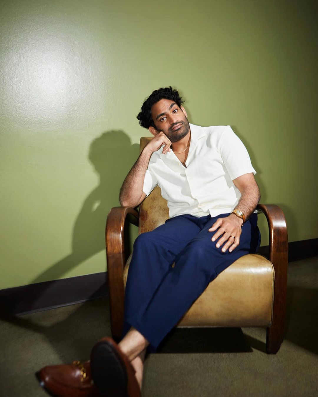 Man in a white shirt and blue trousers seated in a leather armchair against a green wall