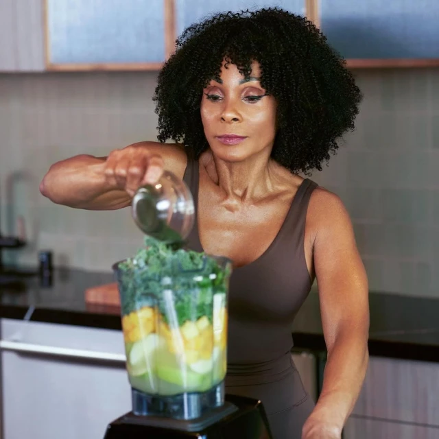 Adult woman in brown athletic wear pours green leafy vegetables from a clear container into a blender filled with fruits in a home kitchen.
