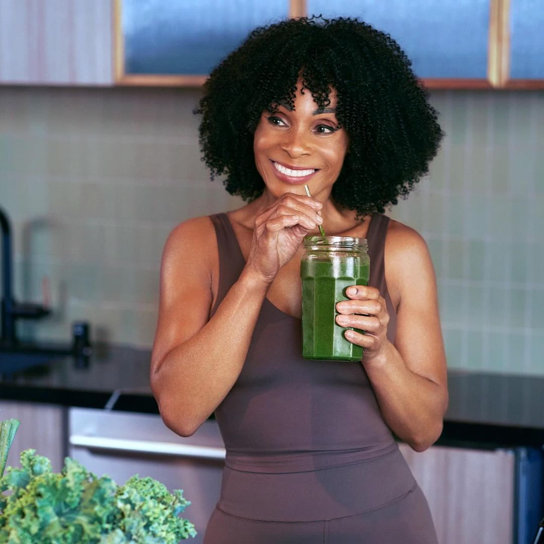 Adult female drinks a green smoothie from a jar with a straw in a home kitchen setting with kale in foreground.