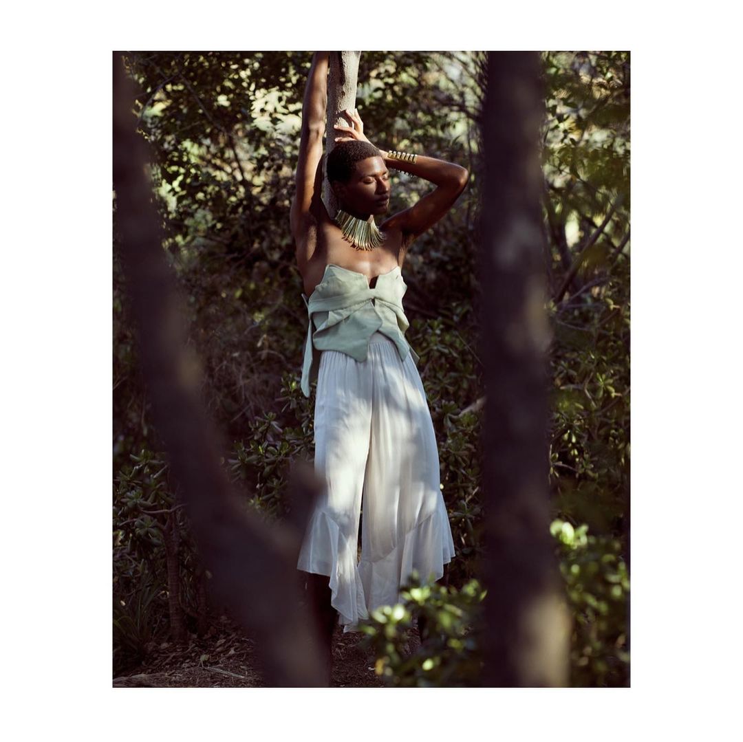 Black model with short hair wearing a strapless light green top and white flowing trousers posing in a forest.