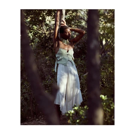 Black model with short hair wearing a strapless light green top and white flowing trousers posing in a forest.