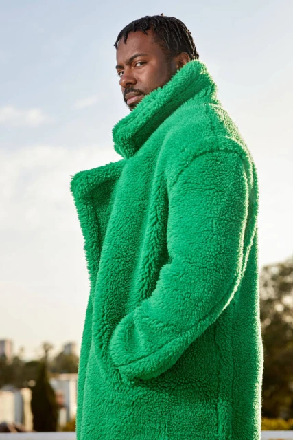 Portrait of a handsome African American man wearing a bright green oversized coat against a blue sky, looking confidently at the camera.