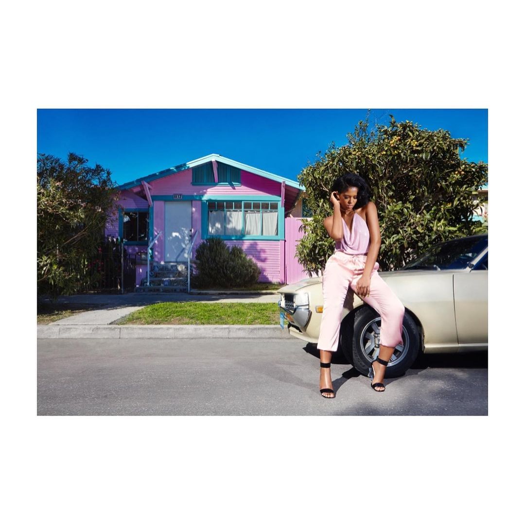 An adult female sits on the hood of a car parked on a street in front of a pink house, wearing a pale pink outfit and black high heels.