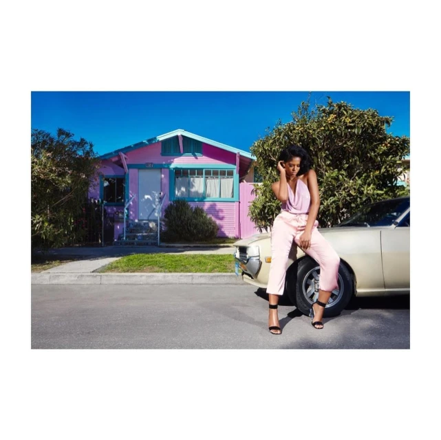 An adult female sits on the hood of a car parked on a street in front of a pink house, wearing a pale pink outfit and black high heels.