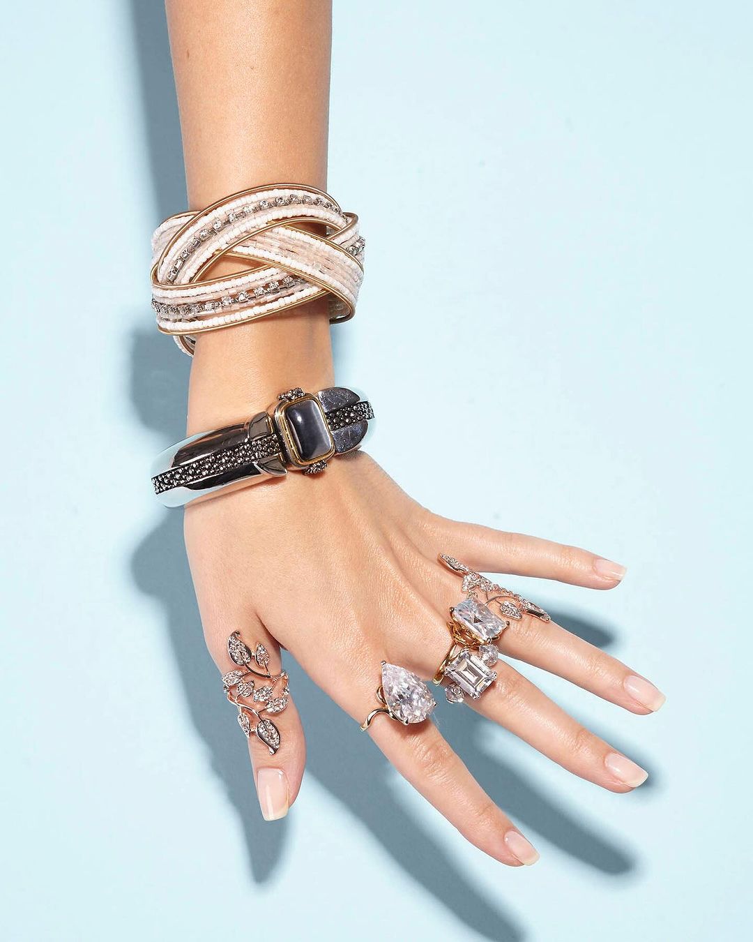 Woman's hand adorned with statement rings and bracelets, featuring intricate designs and gemstone accents against a light blue backdrop.