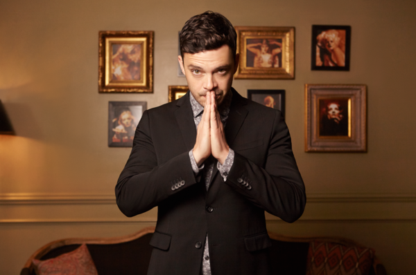 Adult male in a black suit stands indoors with hands together in front of a wall with framed pictures in a medium studio shot.