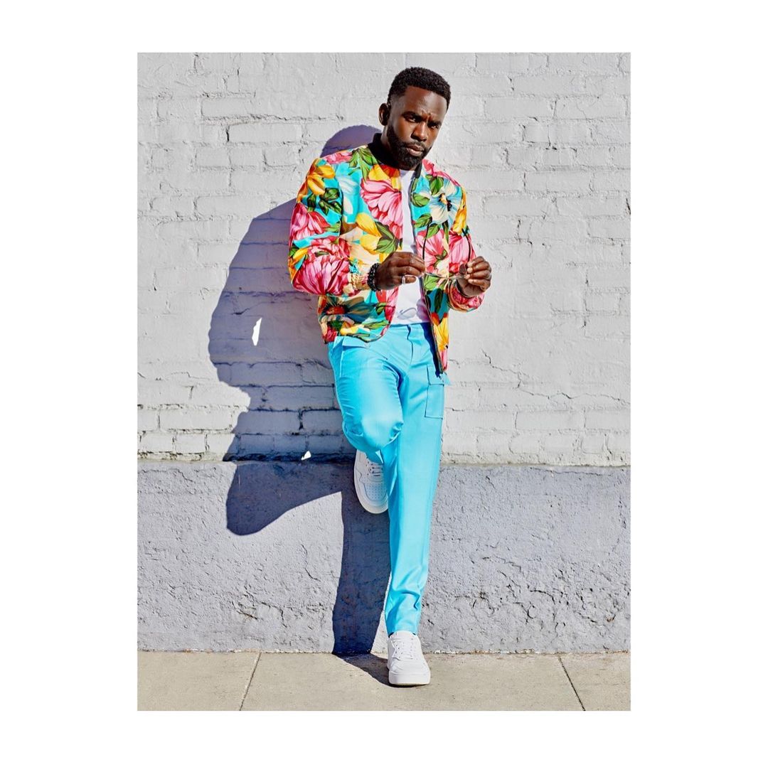 A Black male model is leaning against a textured brick wall in bright sunlight, wearing a floral print bomber jacket and light blue pants with white sneakers.