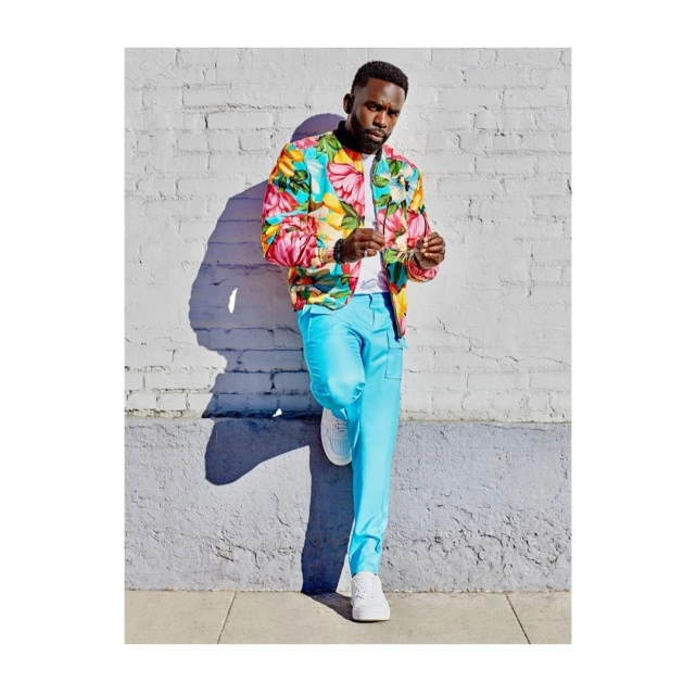 A Black male model is leaning against a textured brick wall in bright sunlight, wearing a floral print bomber jacket and light blue pants with white sneakers.