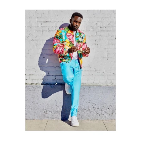 A Black male model is leaning against a textured brick wall in bright sunlight, wearing a floral print bomber jacket and light blue pants with white sneakers.