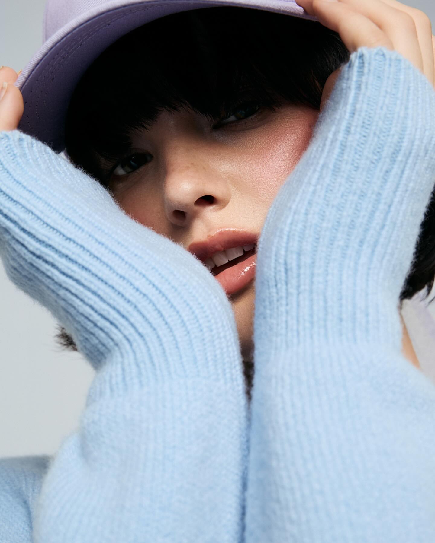 Close-up portrait of a young woman in a light blue sweater and purple baseball cap, with hands framing her face against a neutral background.
