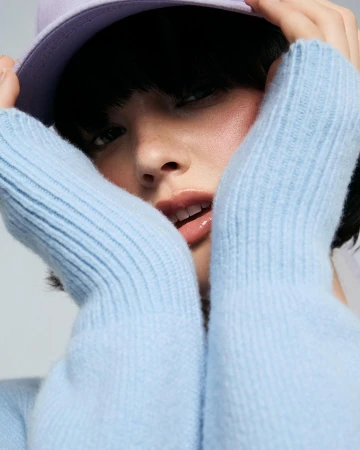 Close-up portrait of a young woman in a light blue sweater and purple baseball cap, with hands framing her face against a neutral background.