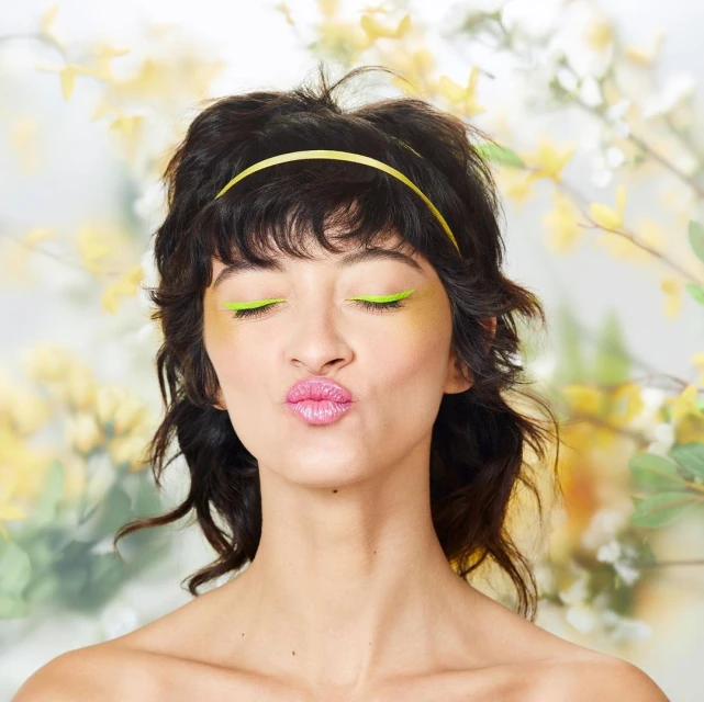 Close-up portrait of a young woman with closed eyes, wearing a yellow headband and bright green eyeliner, against a floral background.