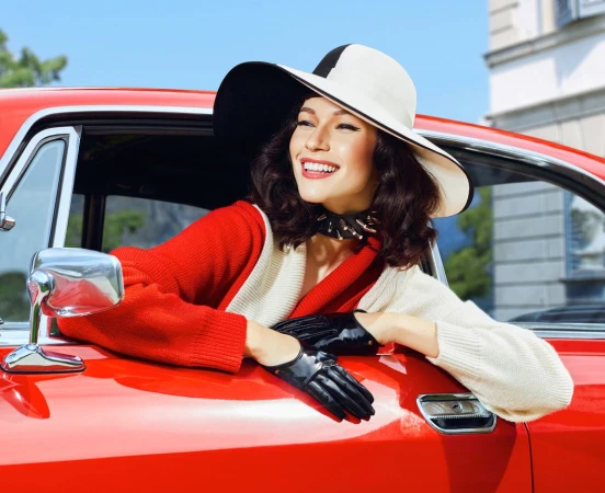 A stylish woman with a wide-brimmed hat leans out the window of a vintage red car, smiling against a backdrop of blue sky and building.