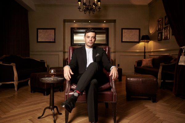 Man in black suit seated in leather chair in upscale interior with glass of whiskey on table. Elegant ambiance, stylish socks.