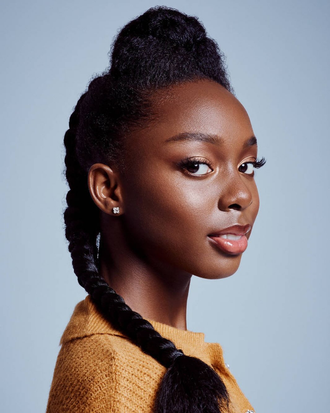 Portrait of a young Black woman with braided hair looking to the side against a light blue background, wearing a mustard yellow sweater.