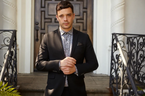 Portrait of a dark-haired man in a black suit with floral shirt, standing outdoors in front of a stone building with a dark wood door.