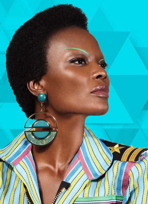 Close-up studio shot of a Black woman with a short afro, wearing statement earrings and a striped jacket against a blue geometric backdrop.