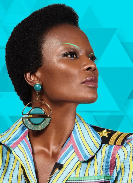 Close-up studio shot of a Black woman with a short afro, wearing statement earrings and a striped jacket against a blue geometric backdrop.