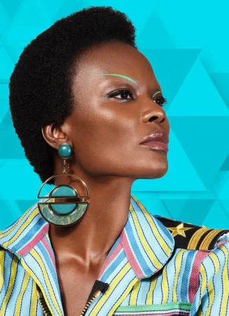 Close-up studio shot of a Black woman with a short afro, wearing statement earrings and a striped jacket against a blue geometric backdrop.