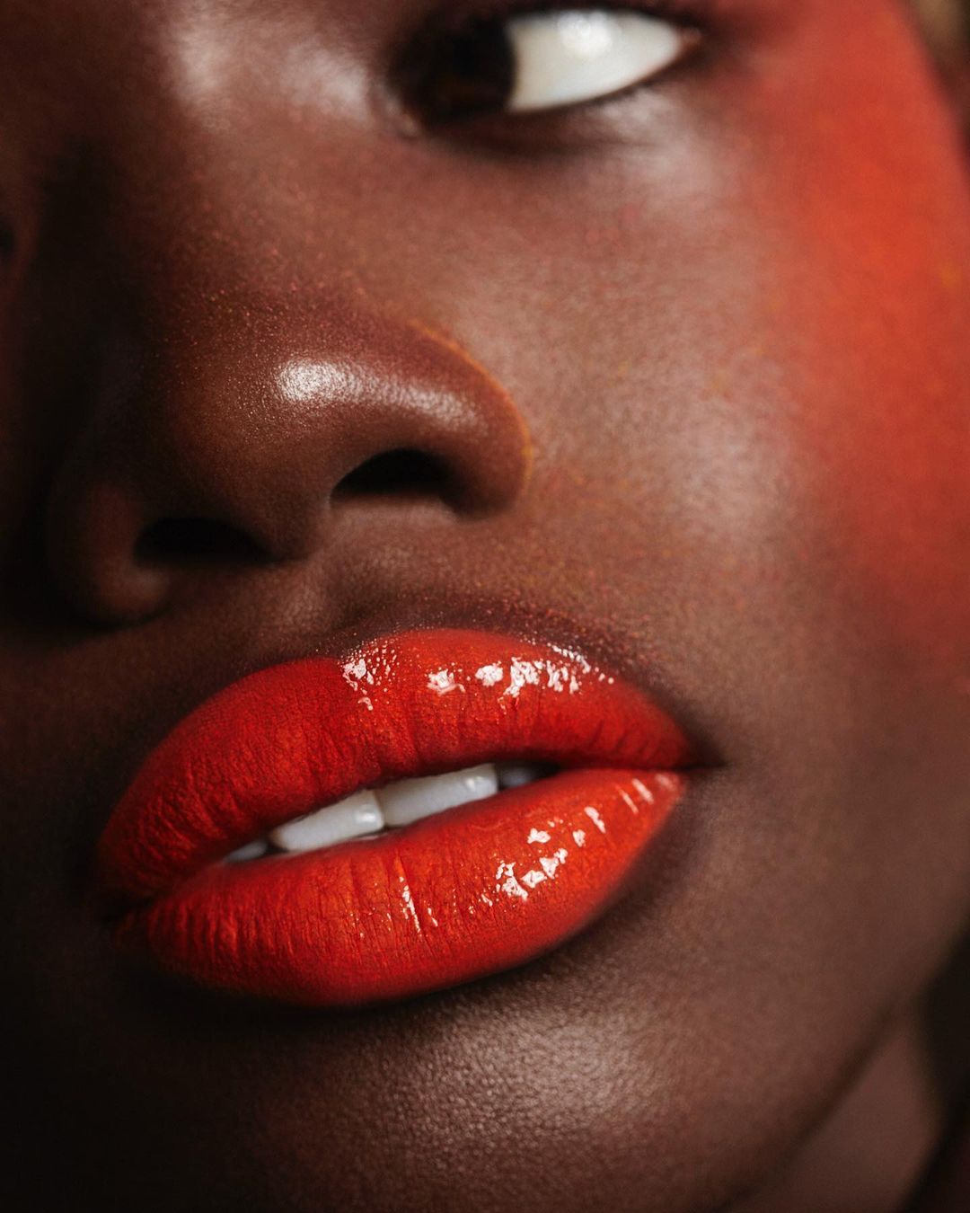 Close-up of a dark-skinned woman's face, highlighting her glossy red lips and subtle blush. Beauty shot showcasing makeup artistry.