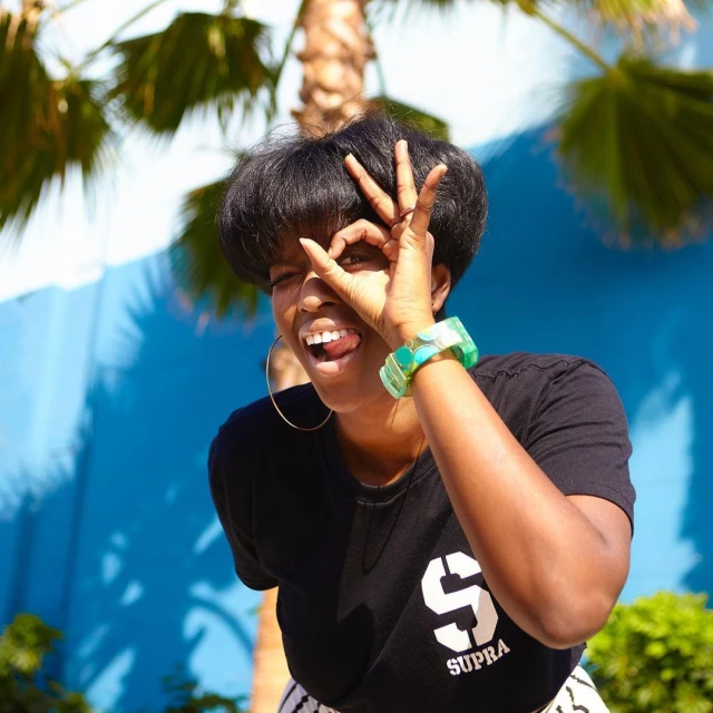 Smiling woman makes an OK hand gesture in front of a blue wall and palm trees, wearing a graphic t-shirt and hoop earrings.