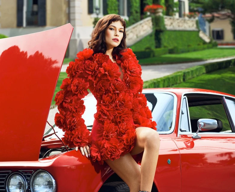 Woman in a red floral dress poses with a vintage red car. The photo features an outdoor setting with a manicured lawn.
