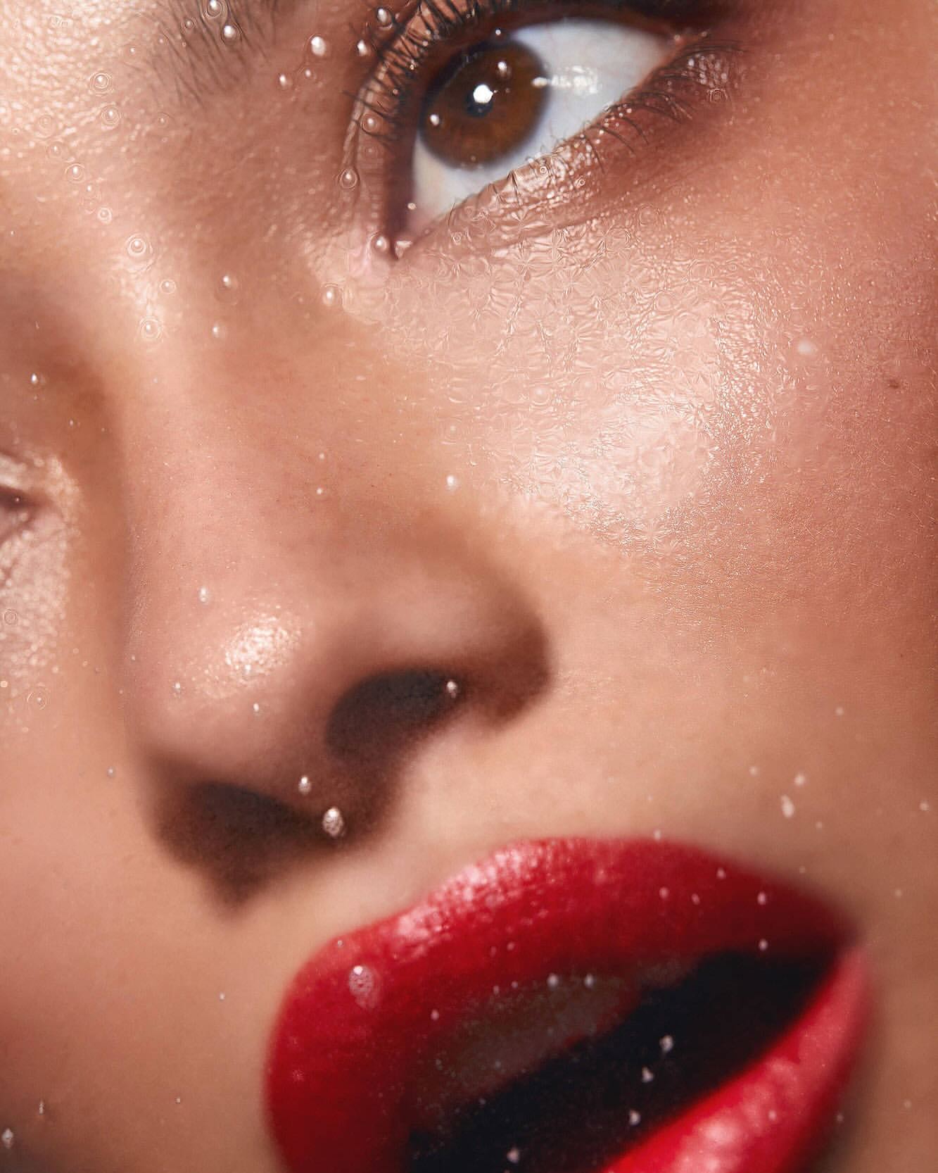 Close-up studio shot of a woman's face, featuring shiny red lipstick and delicate water droplets adorning her skin and around her eye.