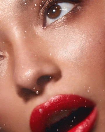 Close-up studio shot of a woman's face, featuring shiny red lipstick and delicate water droplets adorning her skin and around her eye.