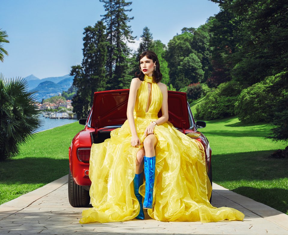 A striking fashion shot featuring a woman in a yellow gown and blue boots sitting on a red car, with a picturesque lake and mountains.