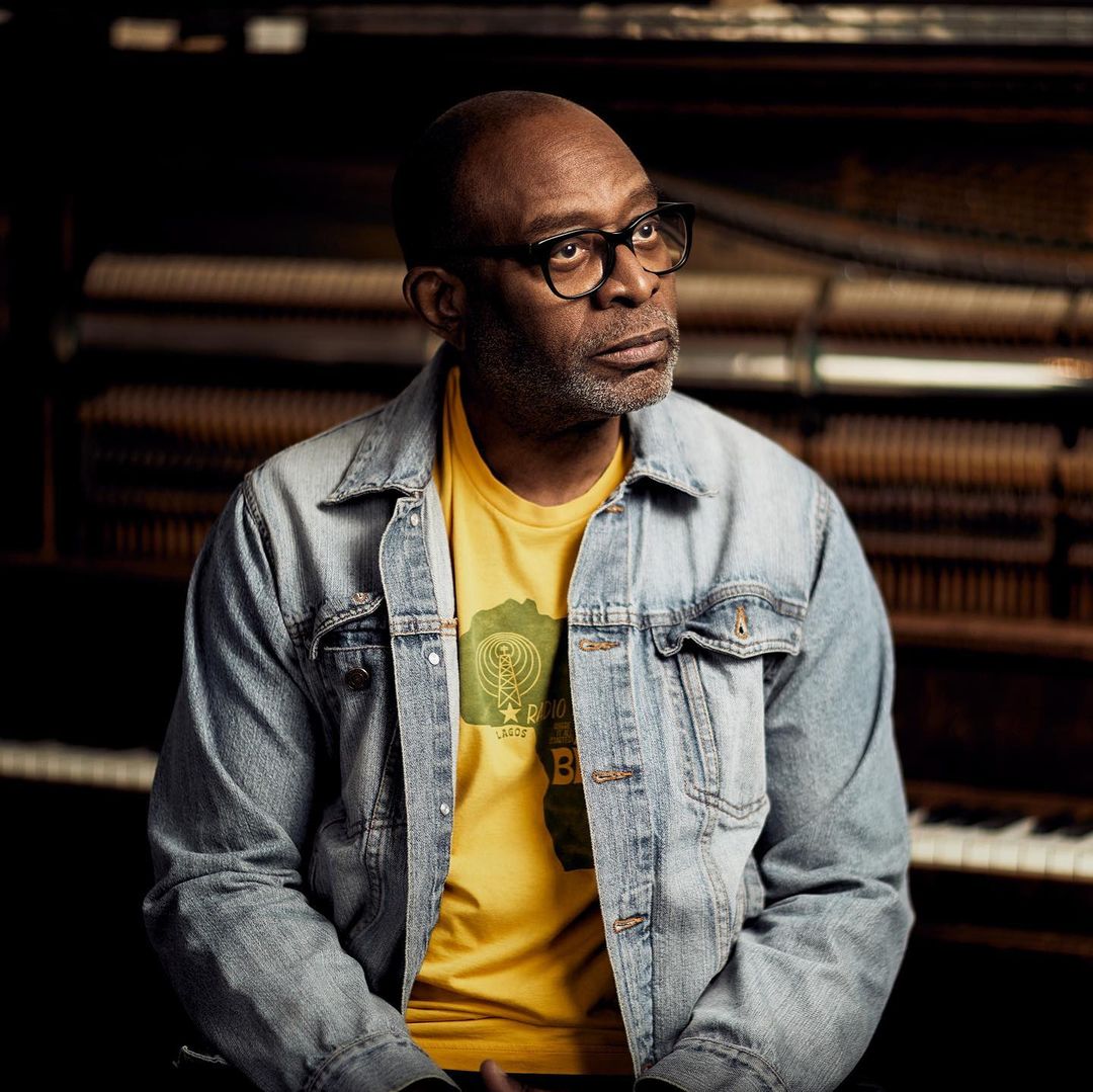 Portrait of a black man with glasses in front of piano, wearing a yellow graphic tee and denim jacket in dim, atmospheric lighting.