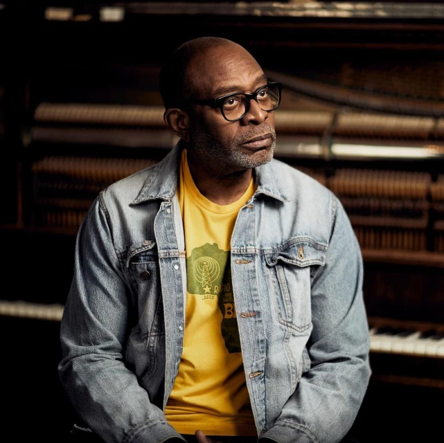 Portrait of a black man with glasses in front of piano, wearing a yellow graphic tee and denim jacket in dim, atmospheric lighting.